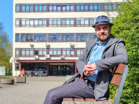 Mężczyzna w kapeluszu (Fifny Przewodnik) siedzi na ławce przed budynkiem Collegium Maximum Uniwersytetu Przyrodniczego w Poznaniu