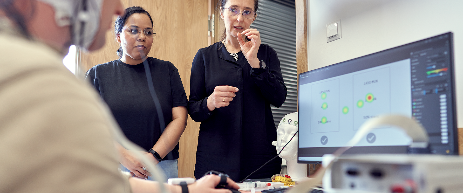 na zdjęciu widać badanie EEG, badany siedzi przy komputerze i patrzy w ekran, dwie badaczki stoją obok niego i wyjaśniają przebieg badania