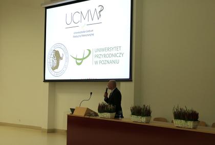 Mężczyzna stoi przed dużym ekranem, wygłaszając prezentację do zgromadzonej publiczności.