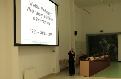 Kobieta stoi przed dużym ekranem, wygłaszając prezentację do zgromadzonej publiczności.