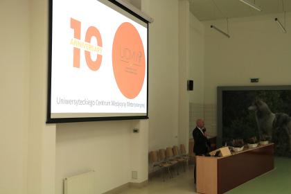 Mężczyzna stoi przed dużym ekranem, wygłaszając prezentację do zgromadzonej publiczności.